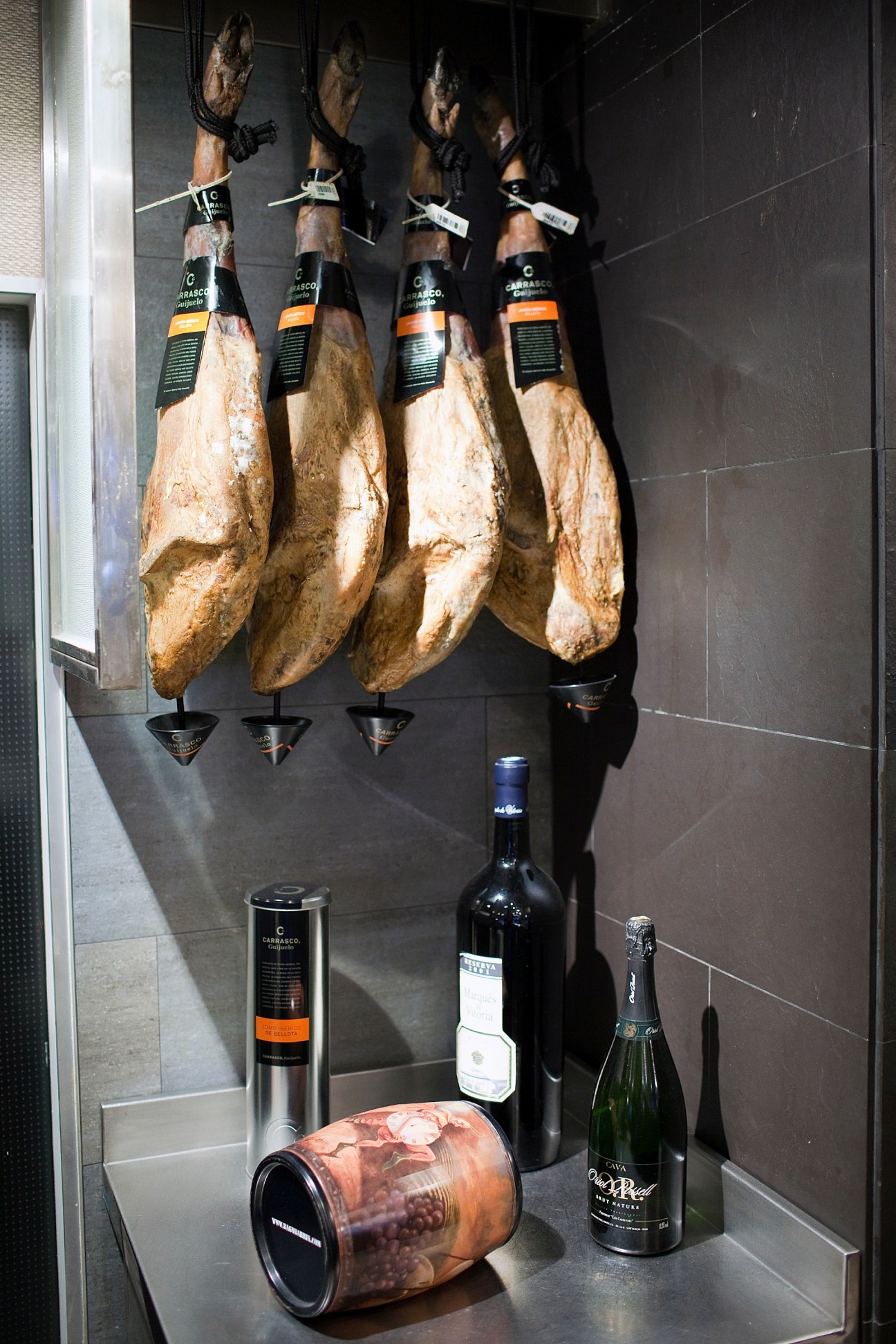 Four cured Jamón Ibérico hams hanging on display with wine bottles and cava below at Andra Mari restaurant.