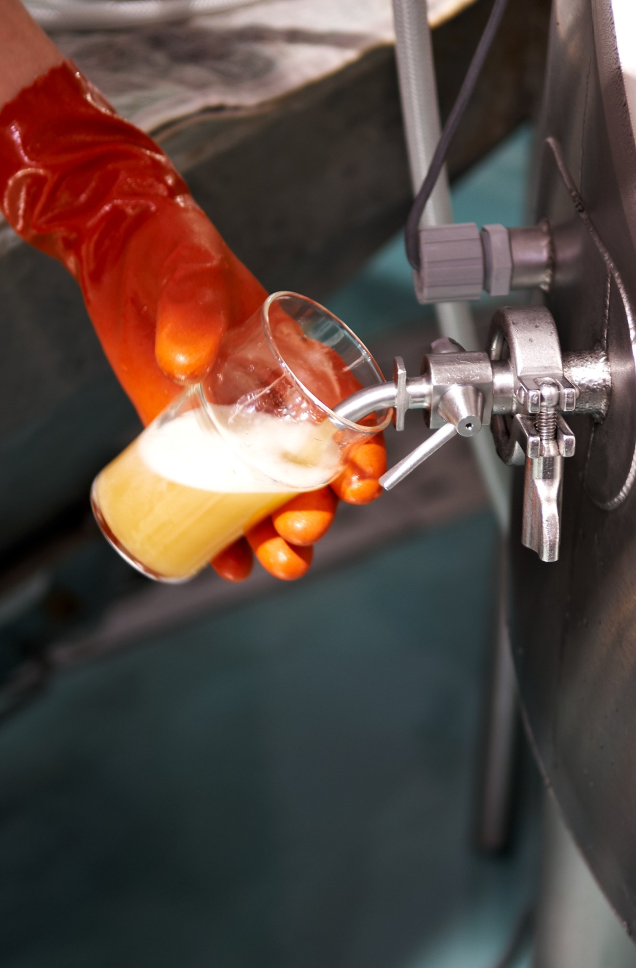 A glass of pale golden beer being poured from a fermentation tank tap, with a red valve visible above.