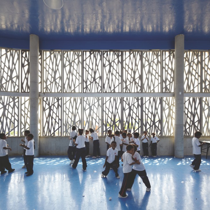 Students in white shirts play in a bright covered courtyard with decorative concrete fretwork panels casting intricate shadows on the blue floor.