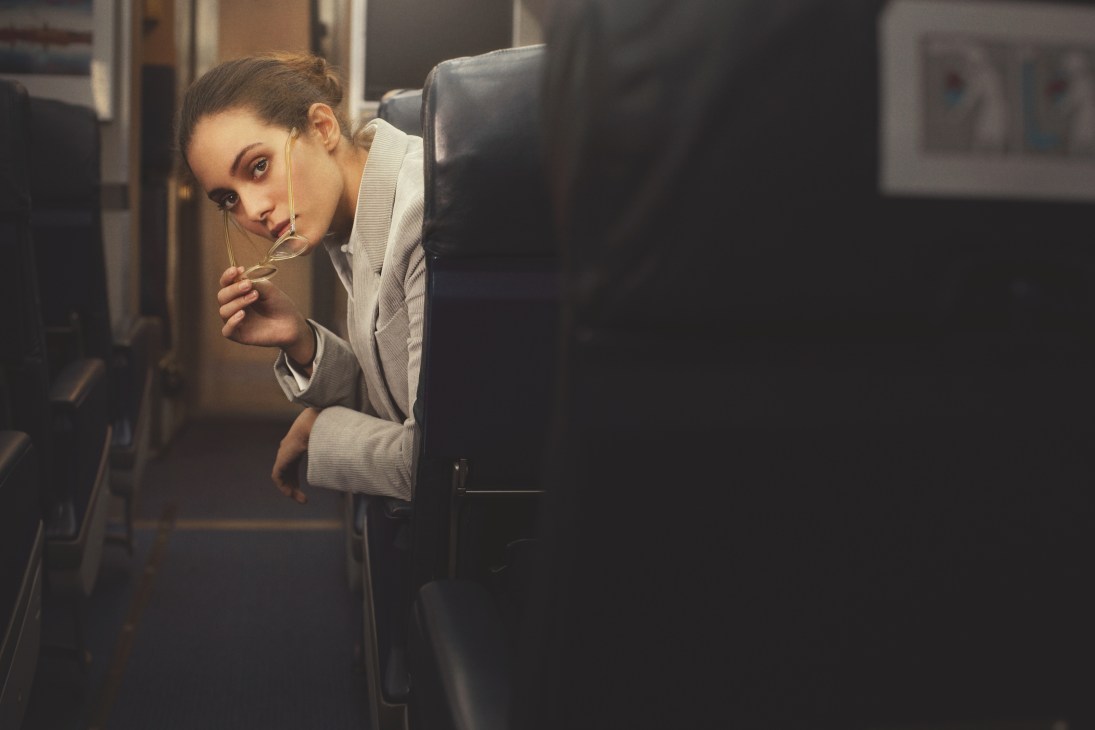 Woman peeking from airplane seat wearing Oliver Peoples glasses, Trussardi jacket and Windsor shirt