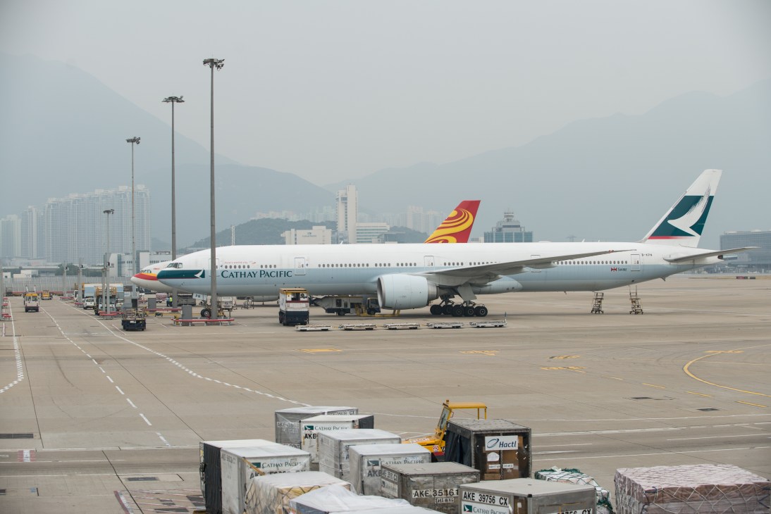 Cathay Pacific Boeing 777-300ER aircraft parked at Hong Kong airport with ground support vehicles and mountainous backdrop.