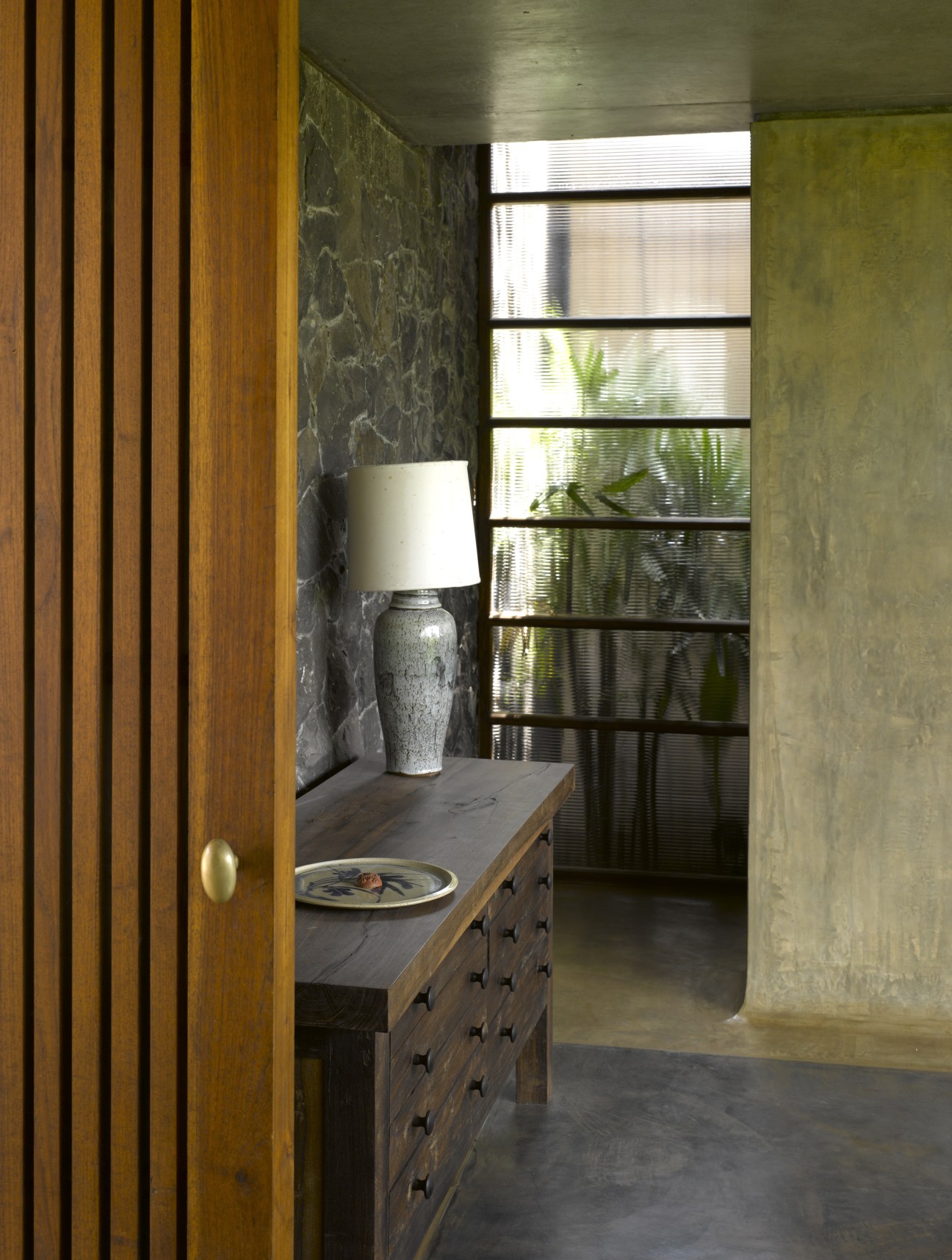 Interior hallway with wooden door, dark stone wall, slatted window screens, wooden chest and ceramic lamp.