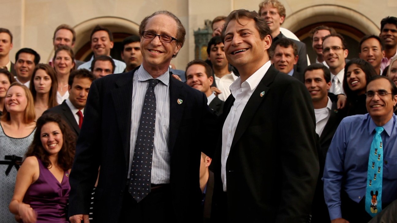 Two men in business attire stand smiling with a large group of people in formal dress behind them against an arched stone building.