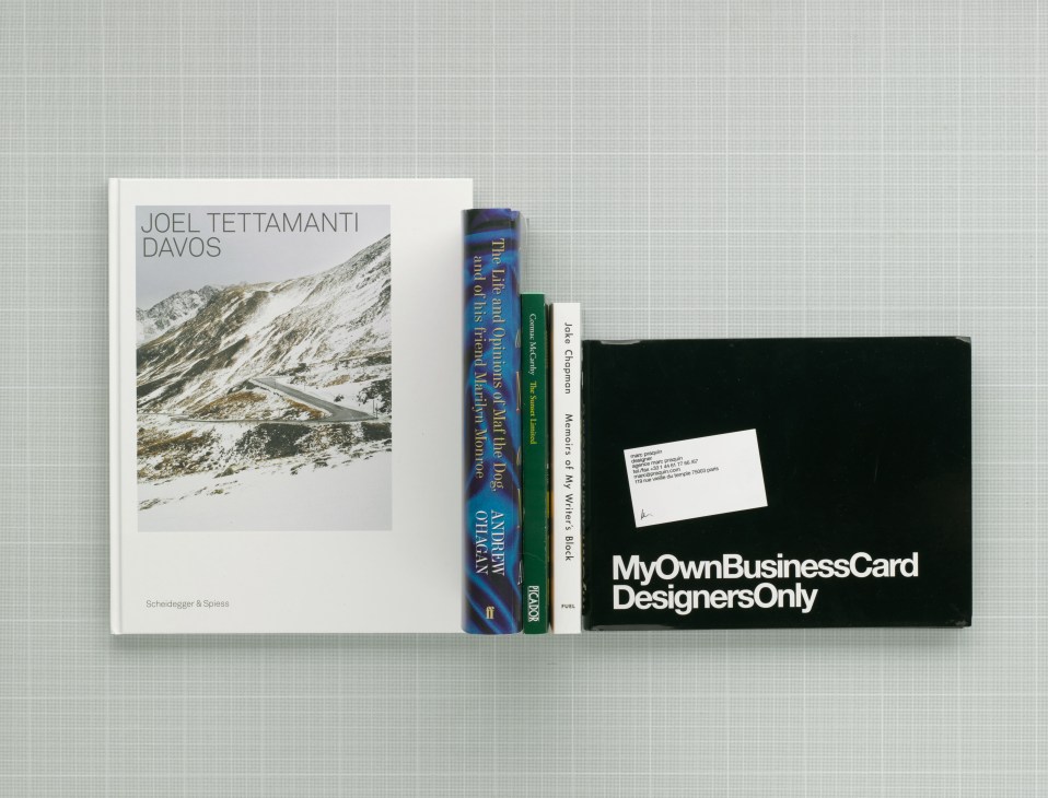 Books displayed on white surface: Joel Tettamanti's Davos photography book, The Life and Opinions of Maf the Dog, The Sunset Limited by Cormac McCarthy, Memoirs of My Writer's Block by Jake Chapman, and MyOwnBusinessCard by Marc Praquin.