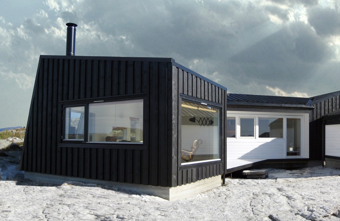 Modern black cabin with white interior sections and large windows on a rocky Norwegian coastal landscape.