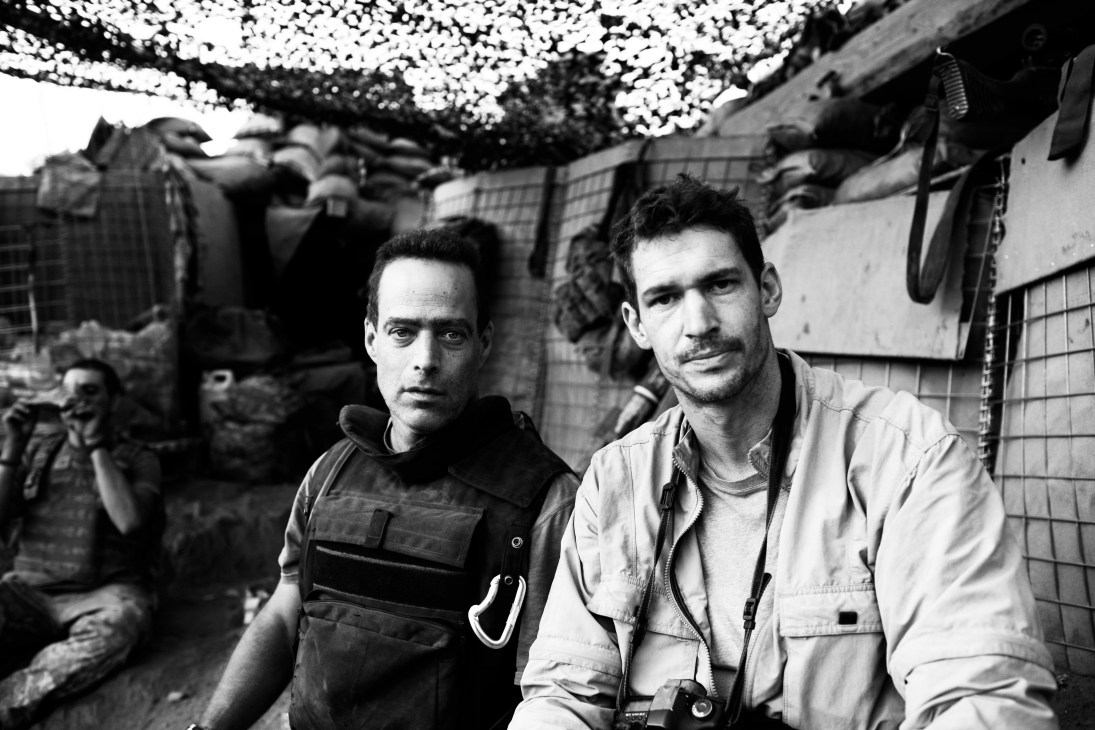 Two men in tactical gear stand in front of military equipment in a war zone setting.