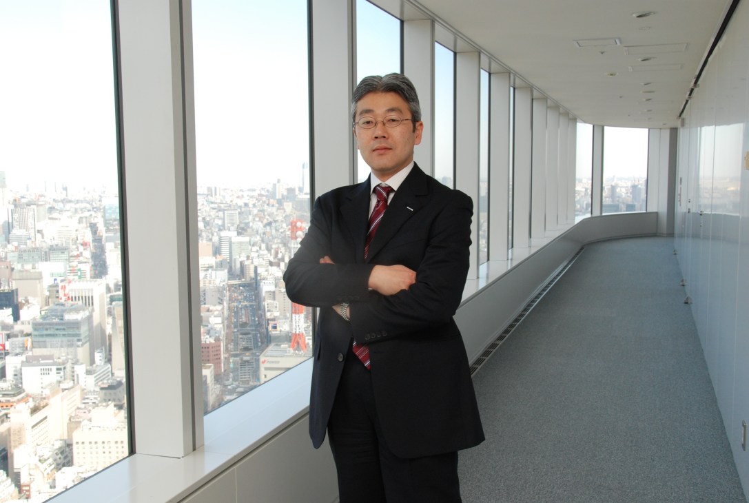 Man in black suit and red striped tie standing in high-rise office with city skyline view.