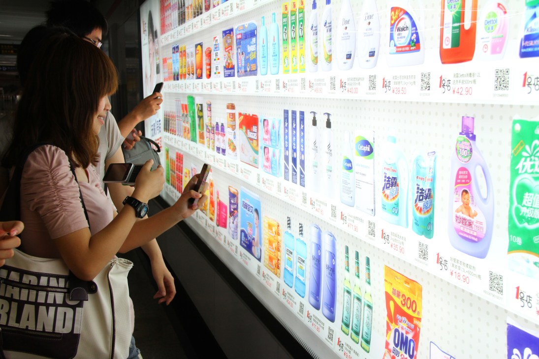 Woman scanning barcode on LED screen displaying products in Shanghai subway virtual supermarket.
