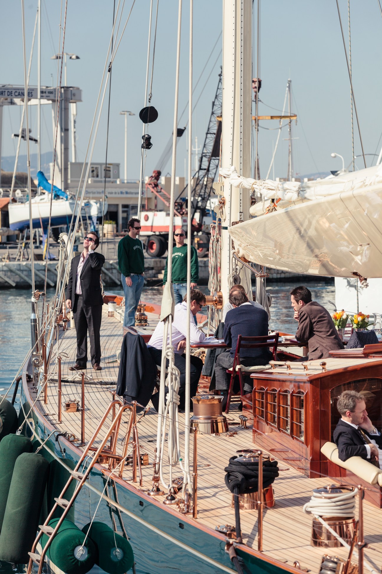 Businesspeople in suits mingling on the deck of a classic sailboat moored in Cannes marina.