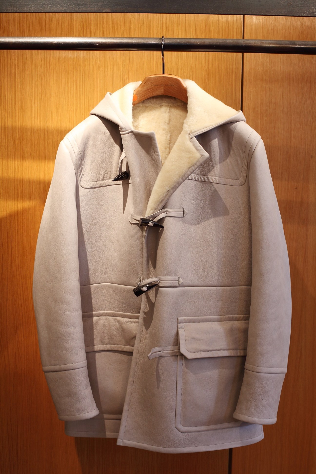 Cream-colored leather jacket with sheepskin lining and toggle button closures displayed on wooden hanger.