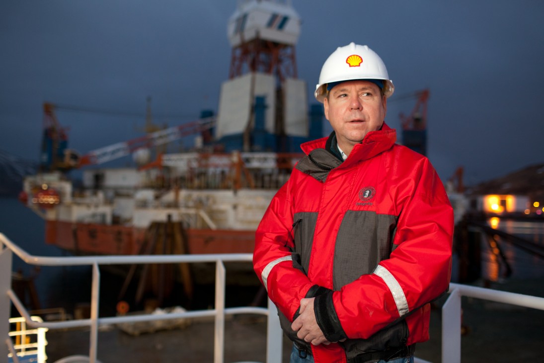 John Kaighin, Shell Oil marine contract manager, wearing a white hard hat and red jacket at Dutch Harbor with industrial port infrastructure in the background.