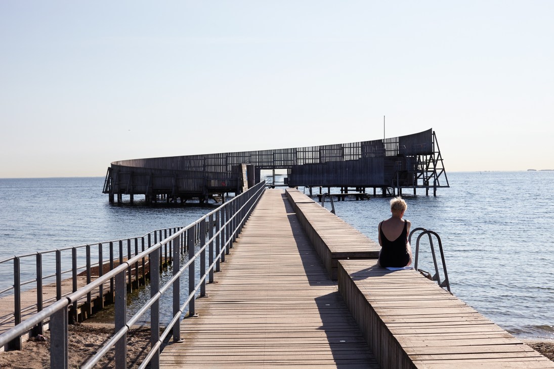 Person sitting on wooden pier extending into calm water with modern pavilion structure in background.