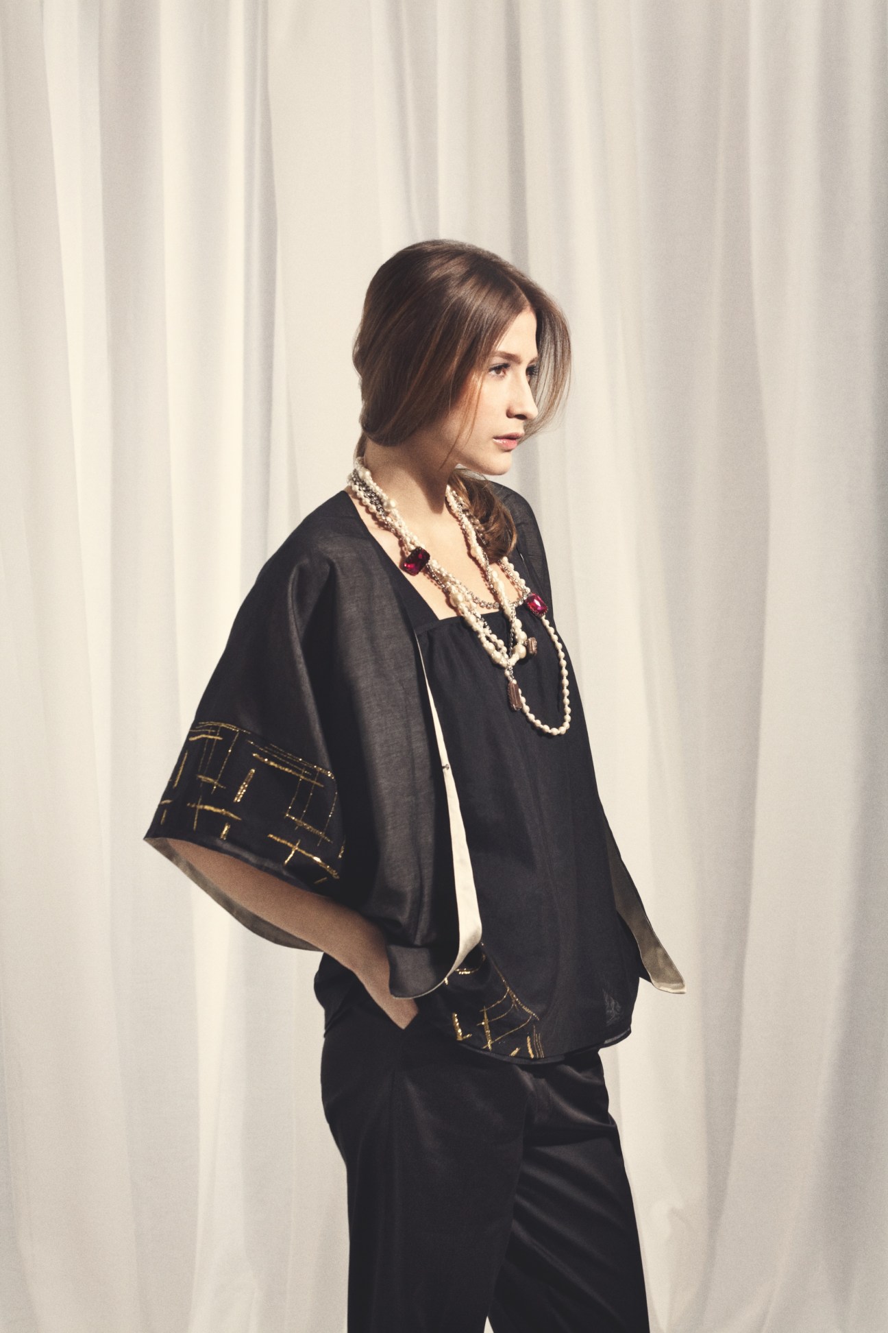 Woman wearing black Duna outfit with embroidered cape and pearl necklaces against white curtain backdrop.