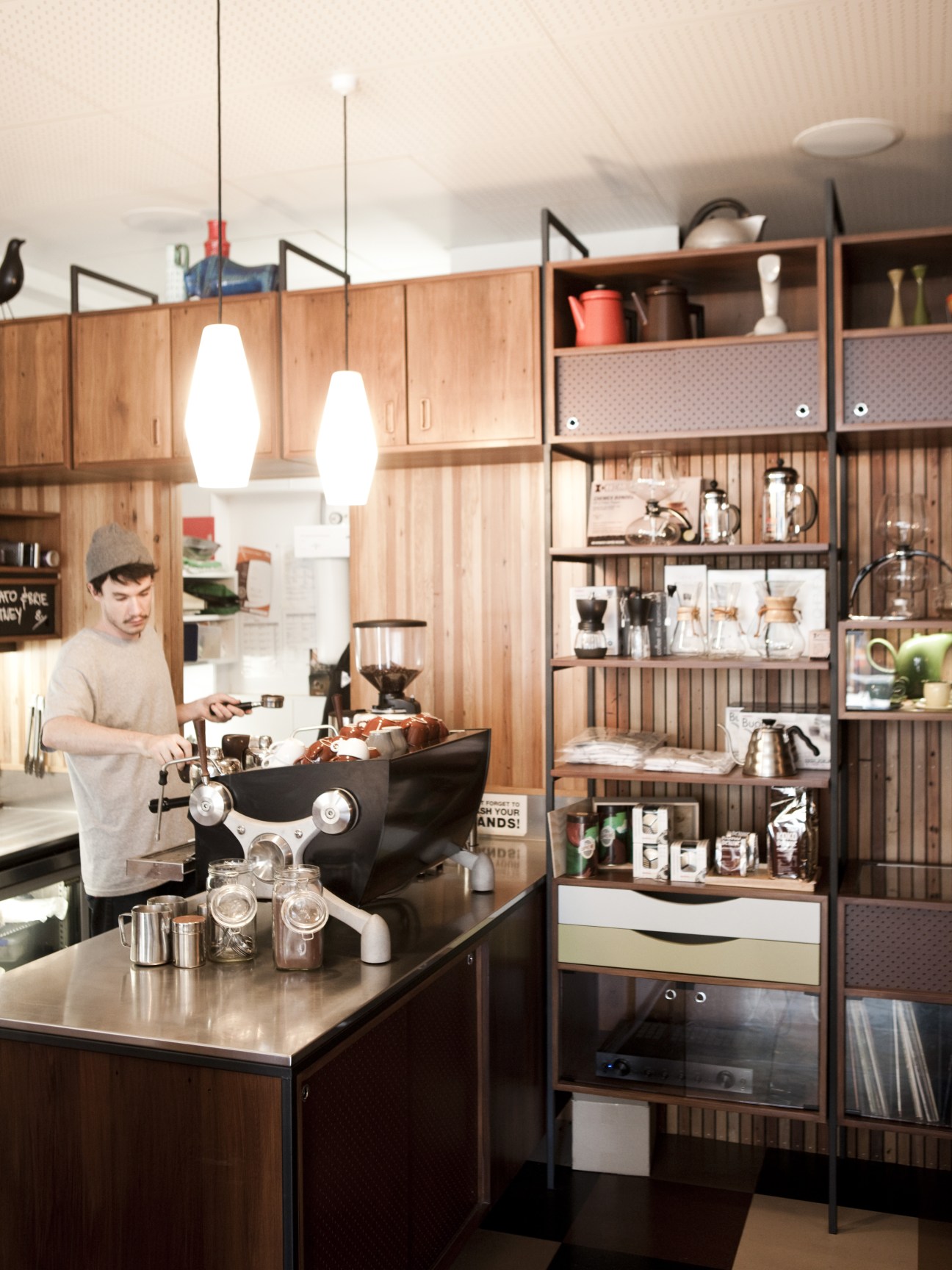 Barista preparing coffee at Customs café in Wellington, with wooden shelving displaying coffee equipment and supplies behind the counter.