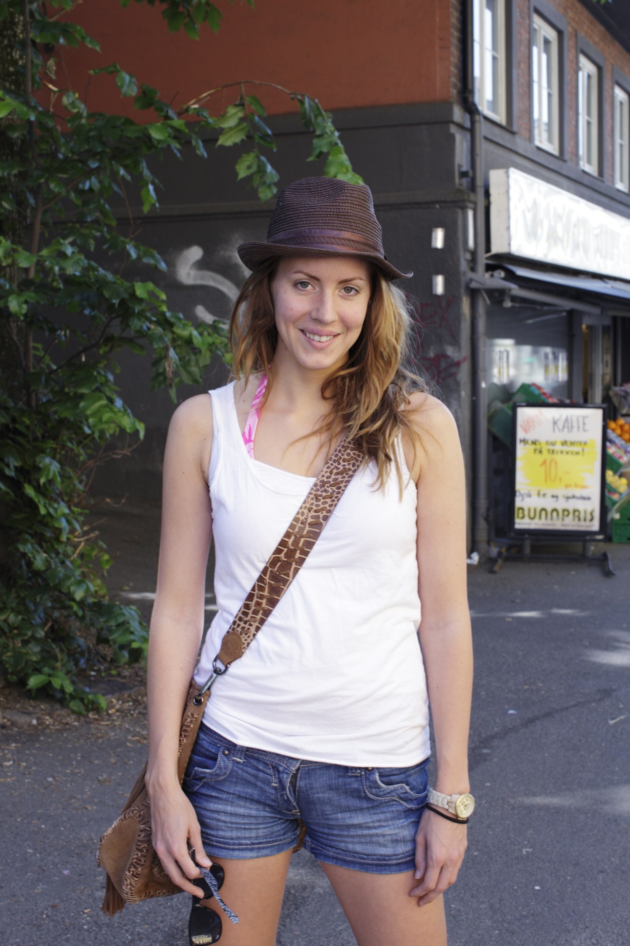 Denise Reitan, a young woman with reddish-brown hair wearing a white tank top, straw hat and patterned shoulder bag, smiling on Thereses gate in Oslo.