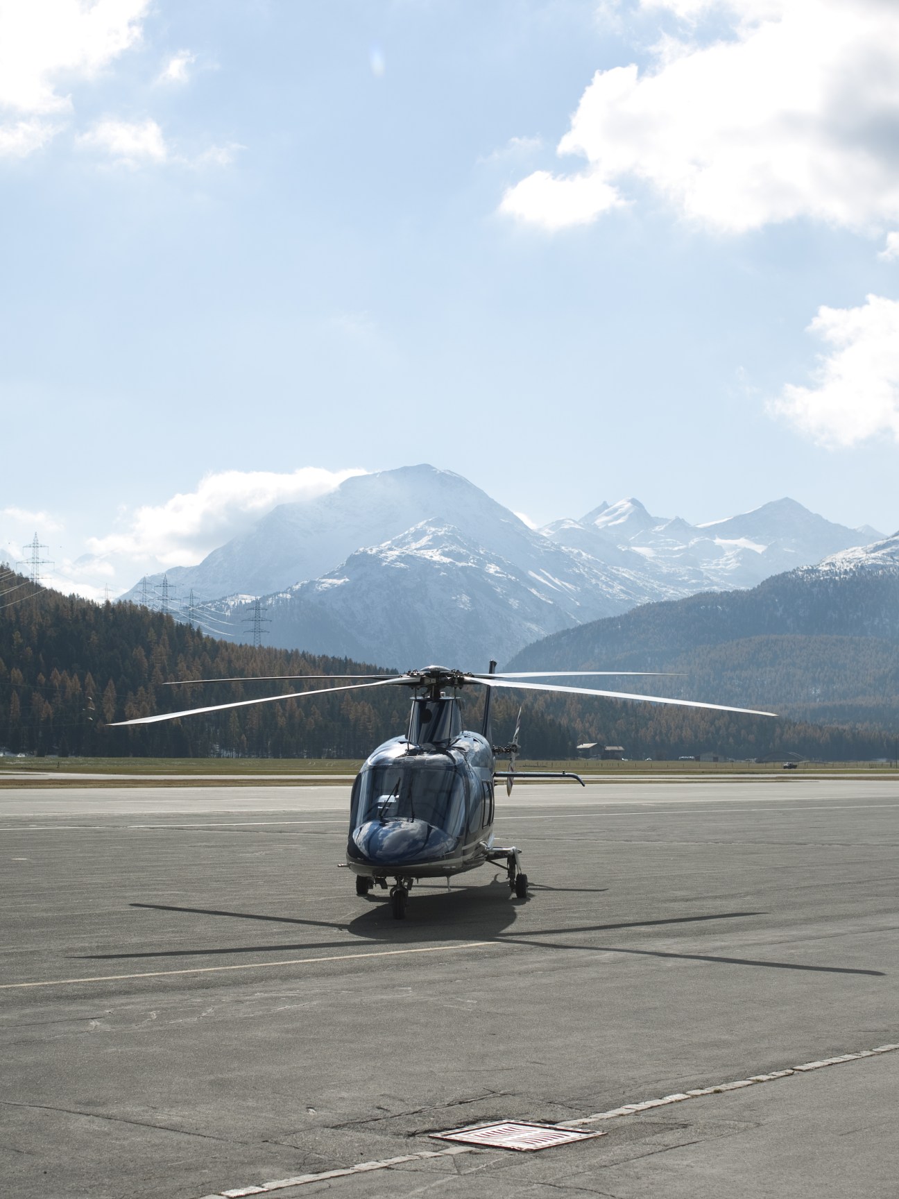 Dark navy helicopter parked on tarmac with snow-capped Alpine mountains in background.