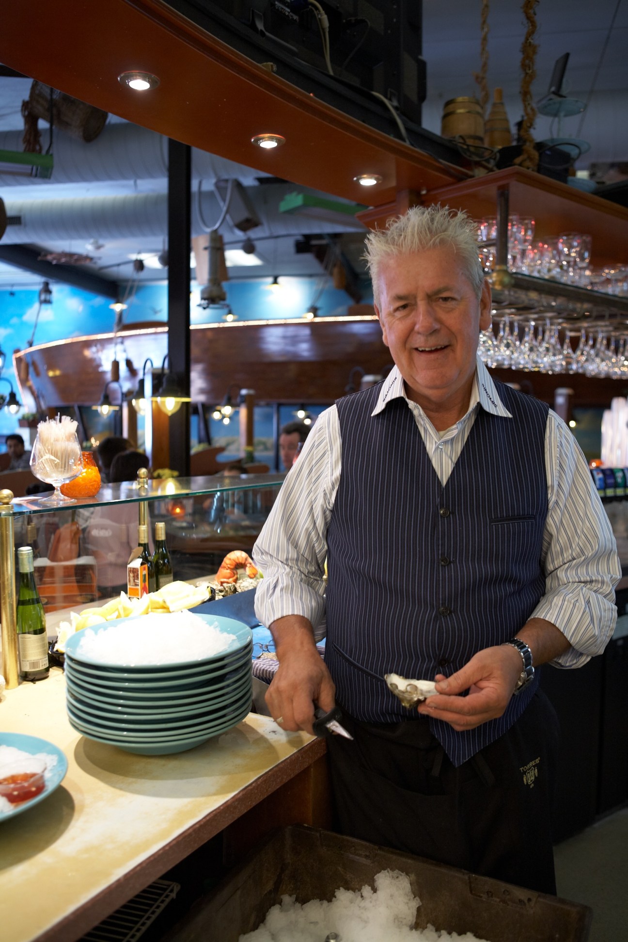 Man at Simonis fish restaurant counter holding an oyster, with plates and glasses nearby.