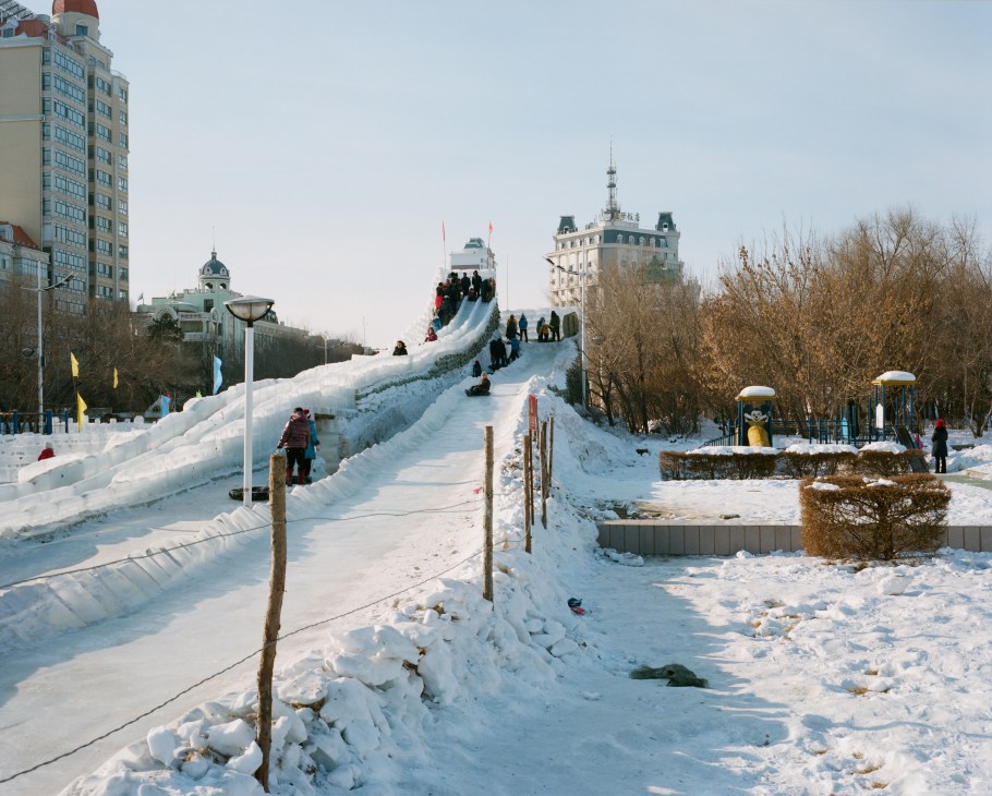 People sliding down a large ice slide in a snowy park with historic buildings visible in the background.