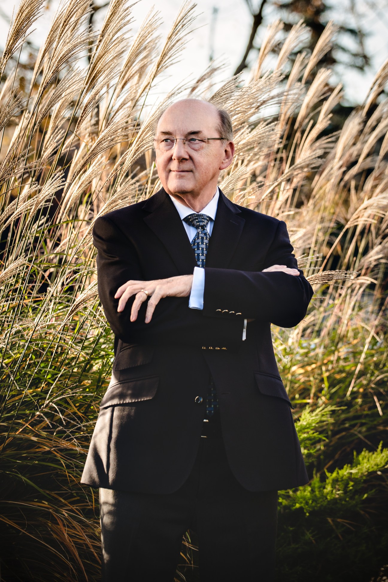 Eddie Friel, museum director, stands in front of tall ornamental grasses wearing a dark suit and glasses.