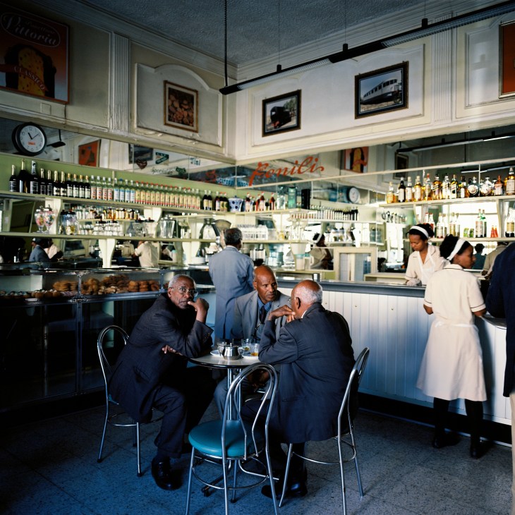 Three men in suits sit at a table inside Valentino Café, a vintage Italian-style diner with white paneling, shelves of bottles, and staff in white uniforms.