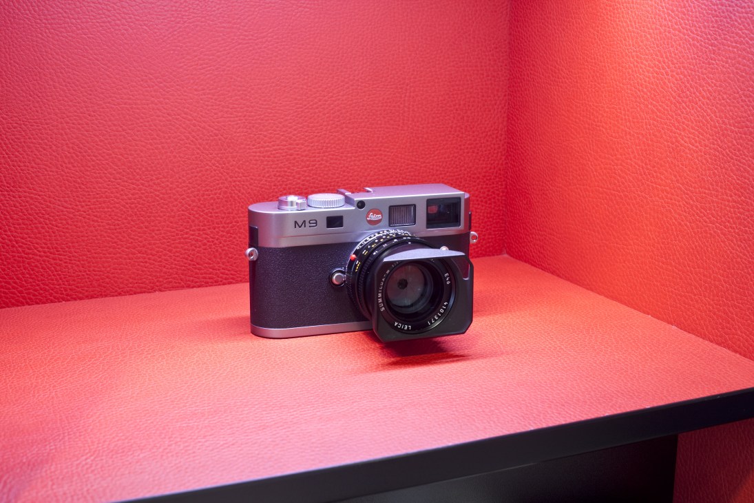Leica M9 rangefinder camera with silver body and black leather grip displayed against pink background.