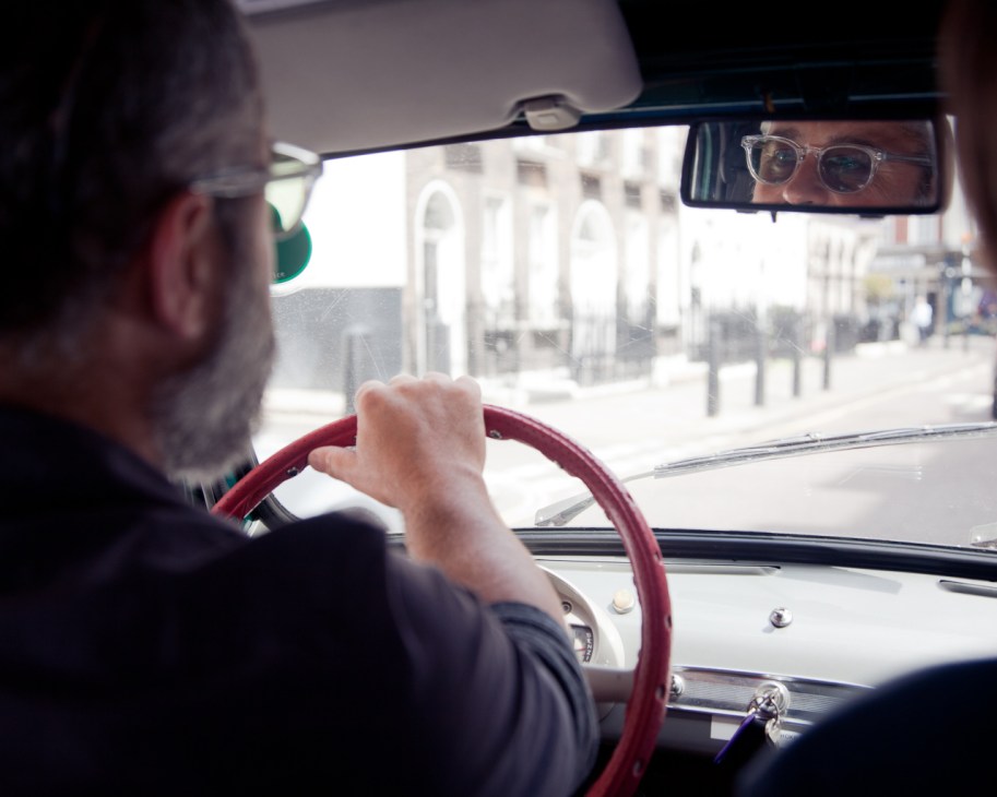 Driver with glasses visible in rear-view mirror, hands on red steering wheel of vintage car.