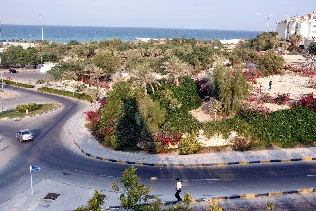 Aerial view of Kish island's landscaped waterfront with palm trees, flowering plants, curved roads, and Persian Gulf beyond.