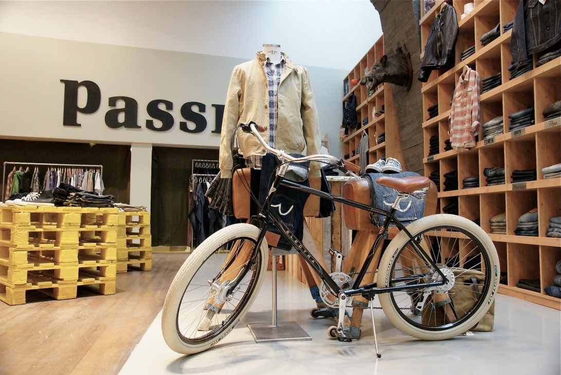 Feldenkirchen retail store interior with mannequin wearing beige jacket over plaid shirt, bicycle with brown leather accessories, wooden shelving, and yellow pallets.