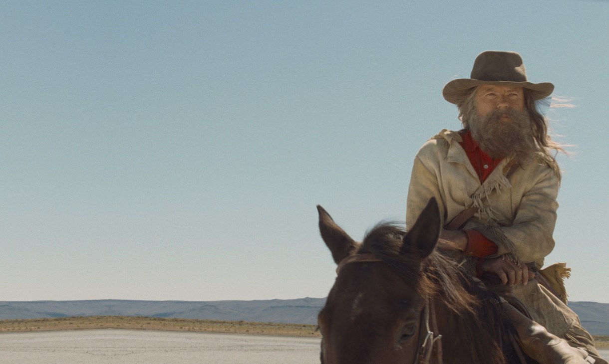 Bearded man in cowboy hat and tan jacket riding horse across desert landscape in Kelly Reichardt's Meek's Cutoff.