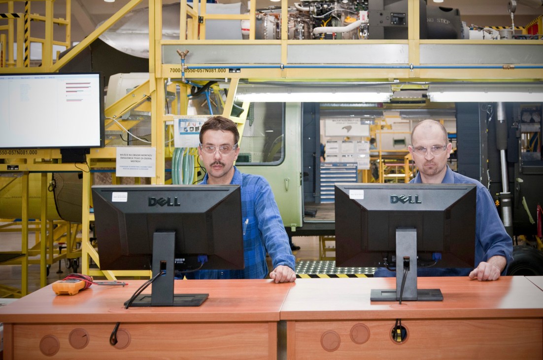 Two male employees in blue work shirts at computer workstations in an aerospace factory with yellow machinery and aircraft components visible in the background.