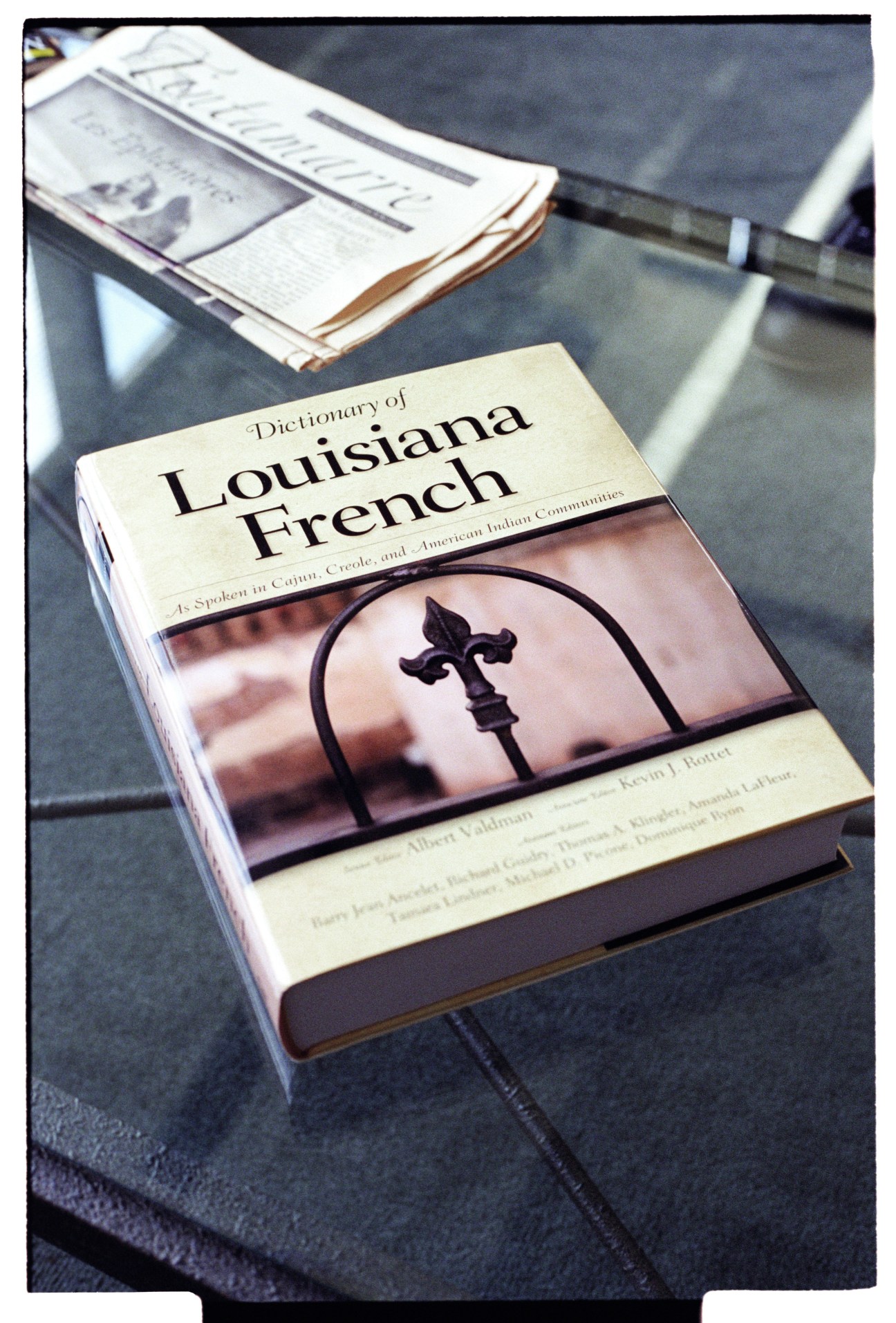 Dictionary of Louisiana French book with fleur-de-lis symbol on cover, displayed on dark surface with newspapers.