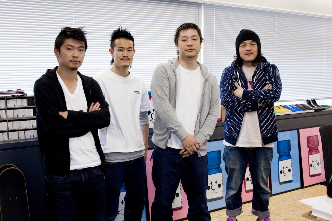 Four male members of the Zozotown web team pose in an office with blinds and colorful boxes behind them.