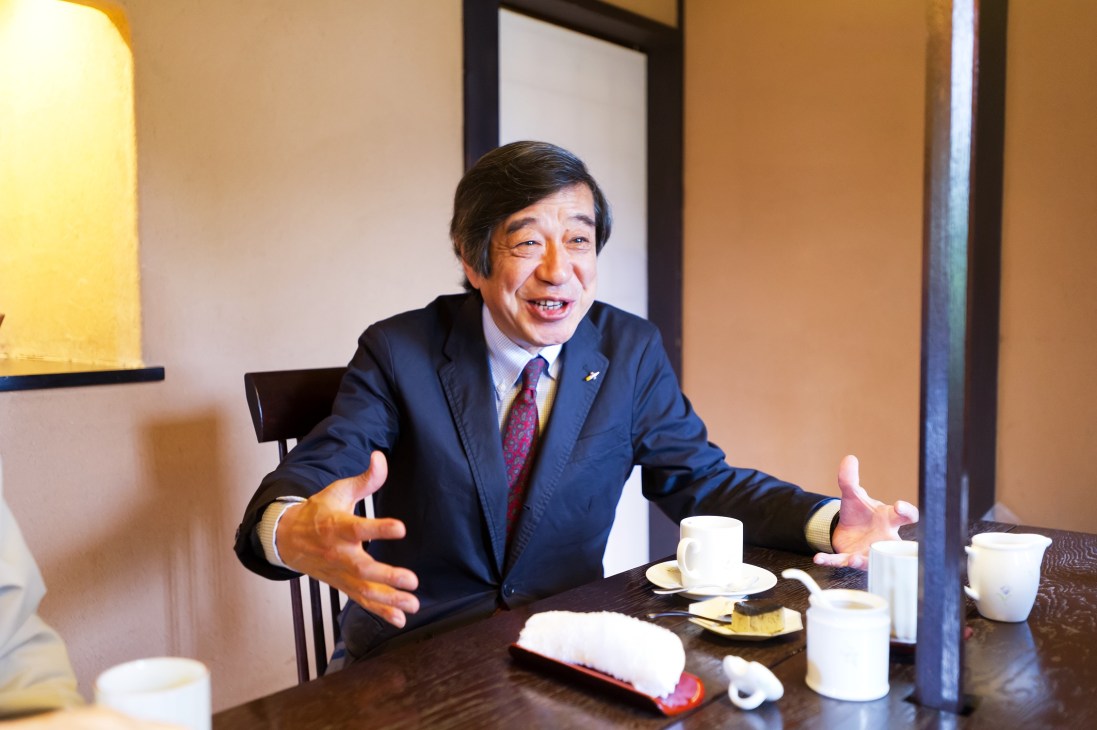 Man in dark suit and red tie gestures while speaking at a table with cups during a meeting.