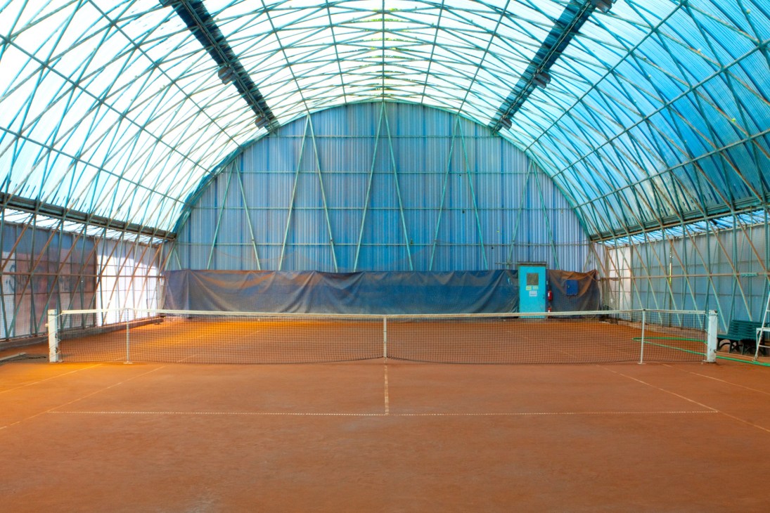 Indoor tennis court with clay surface under a modern glass and metal barrel-vault ceiling structure.