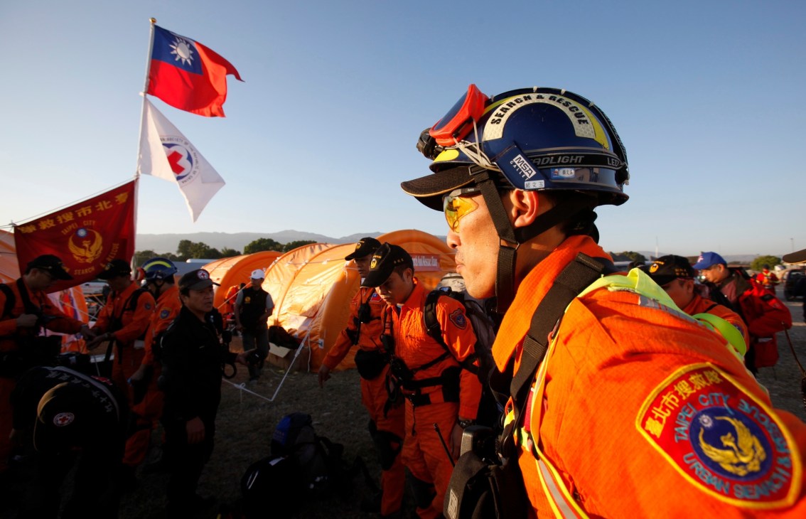 Taiwan rescue workers in orange suits and helmets at a disaster relief camp with Taiwan and search-and-rescue flags.