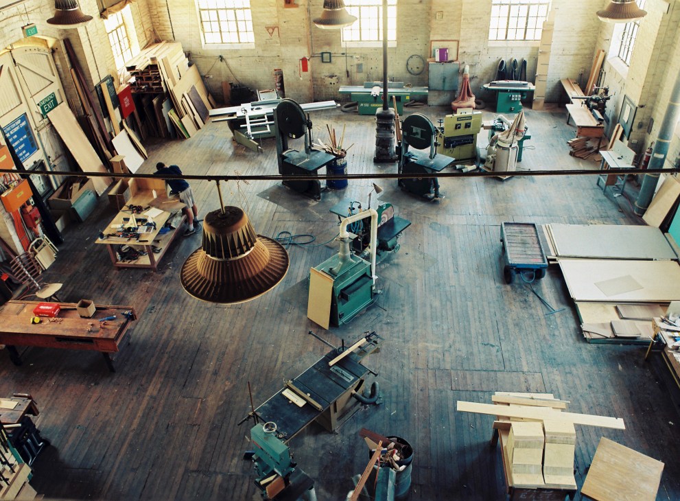 Overhead view of The Pattern Shop workshop showing woodworking equipment, materials, and furniture designers at work in an industrial space.