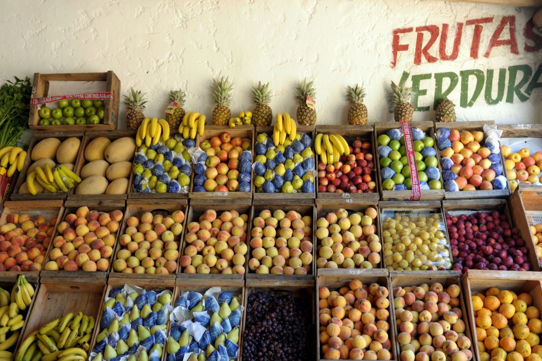 Wooden crates display fresh fruits and vegetables including bananas, pineapples, apples, and oranges at El Tomate Alegre market stall.