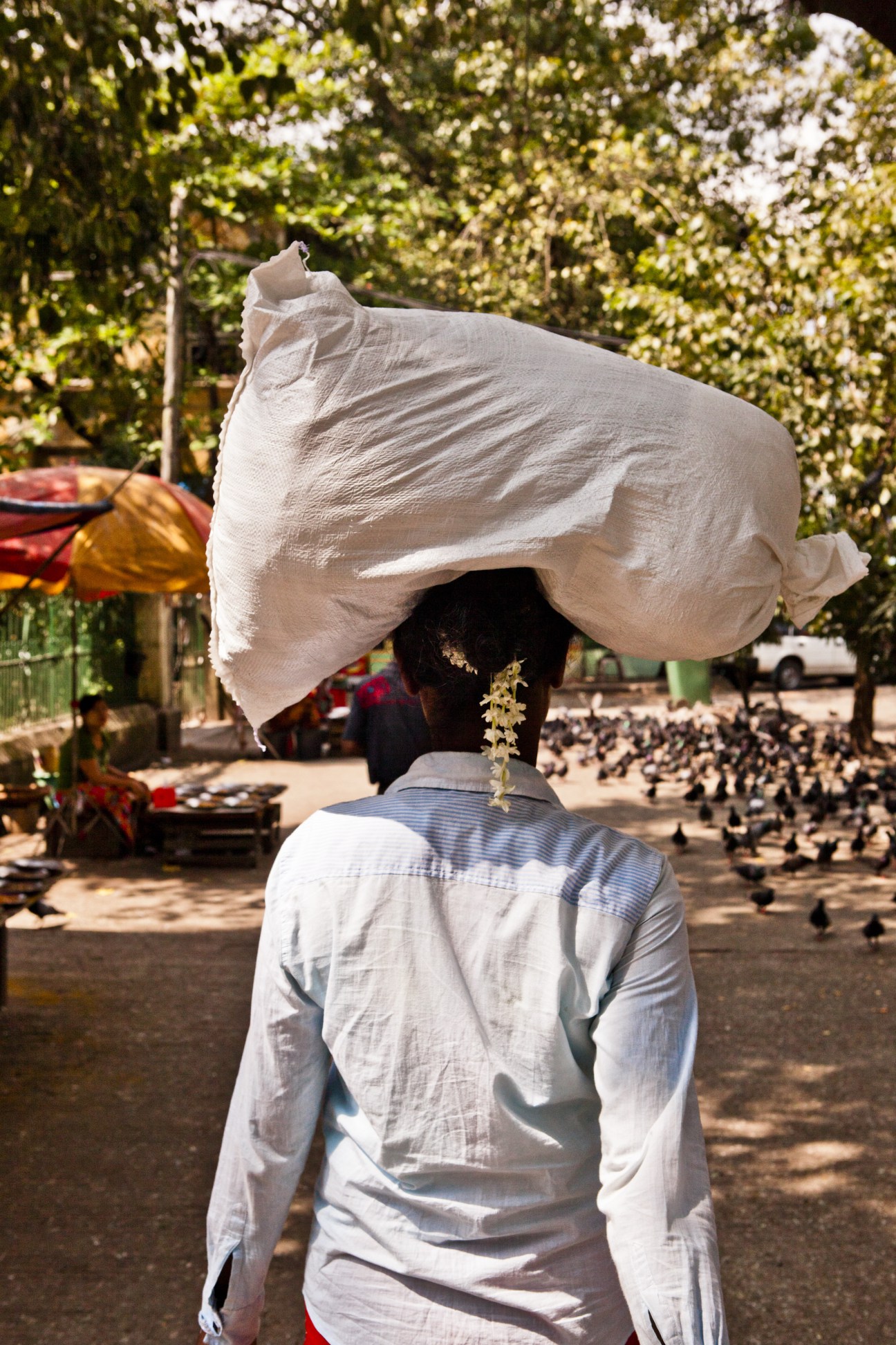 A woman from behind carrying a large white bundle on her head in a sunny public square.