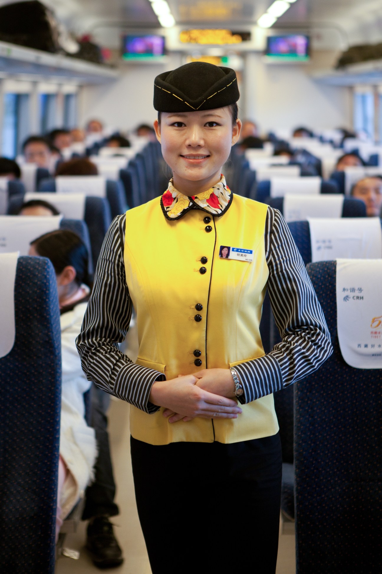 Stewardess in yellow vest and black cap smiling in high-speed train cabin.