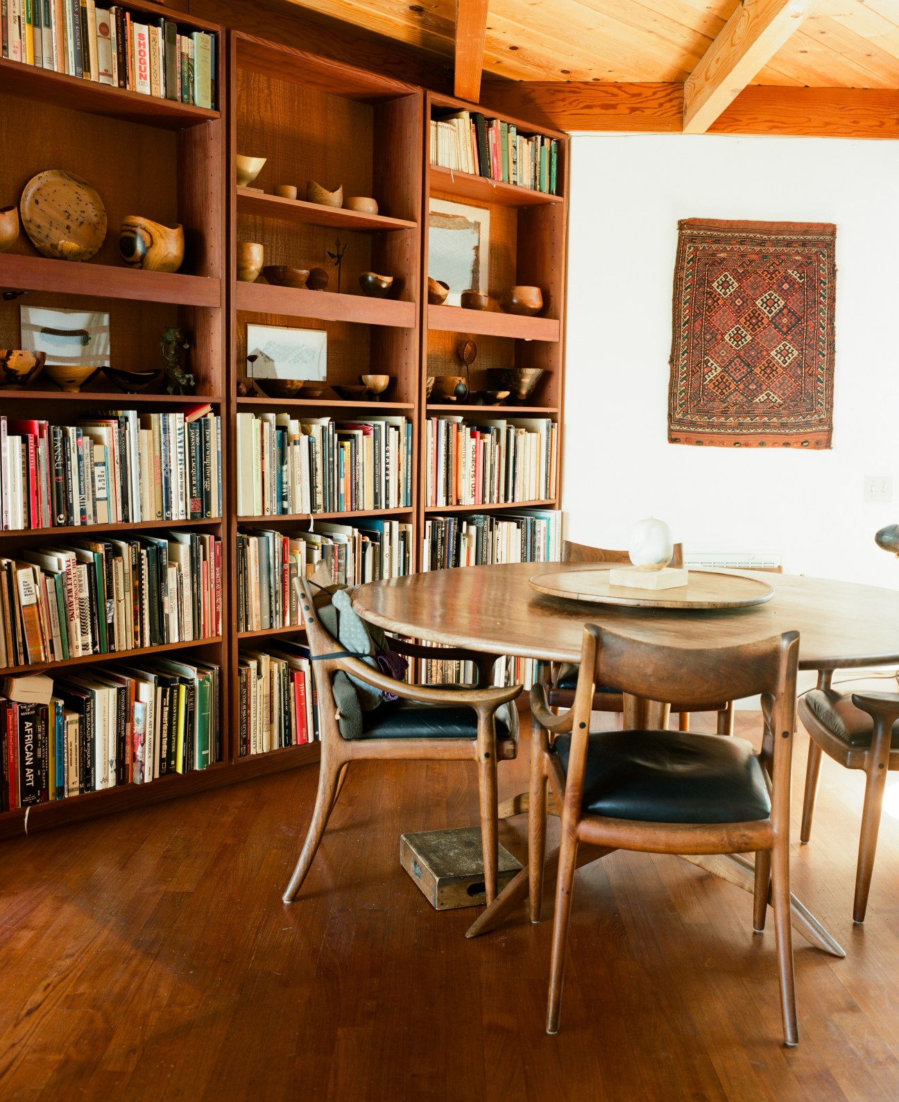 Dining room with wooden table and chairs, built-in bookshelves, woven textile on wall, and exposed ceiling beams.