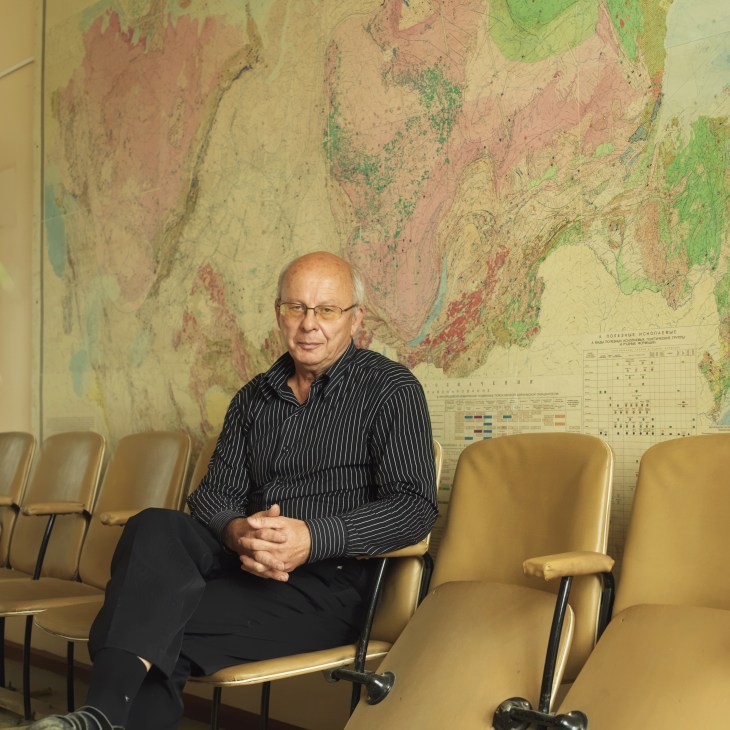 Viktor Okrugin, a volcanologist wearing glasses and a pinstriped shirt, sits in a gold chair in front of a large yellowed geological map of Russia on the wall.