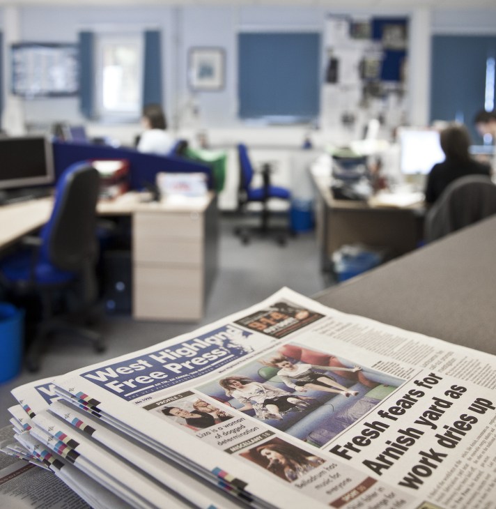 Stacked copies of West Highland Free Press newspaper on desk in newsroom with staff working in background.