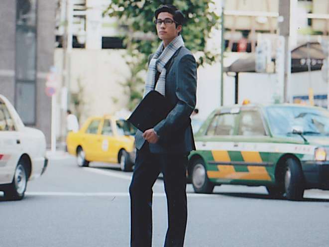 Man in business attire holding a document case stands on a city street with taxis in the background.