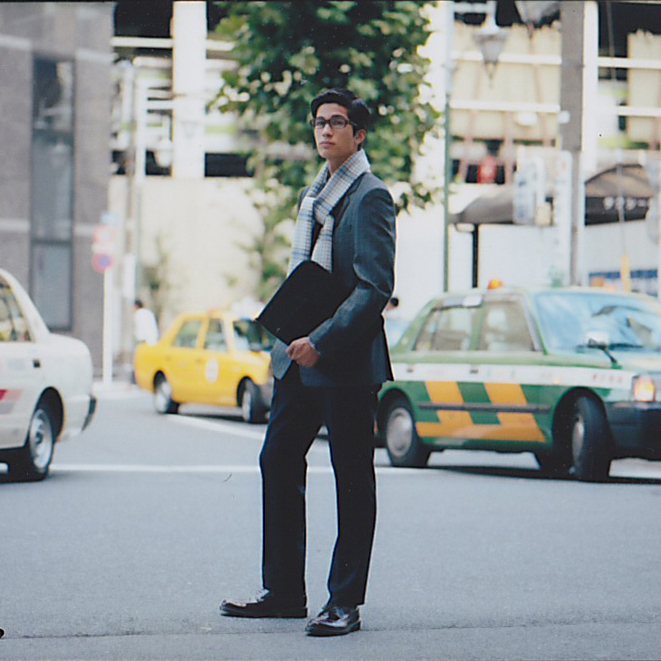 Man in business attire holding a document case stands on a city street with taxis in the background.