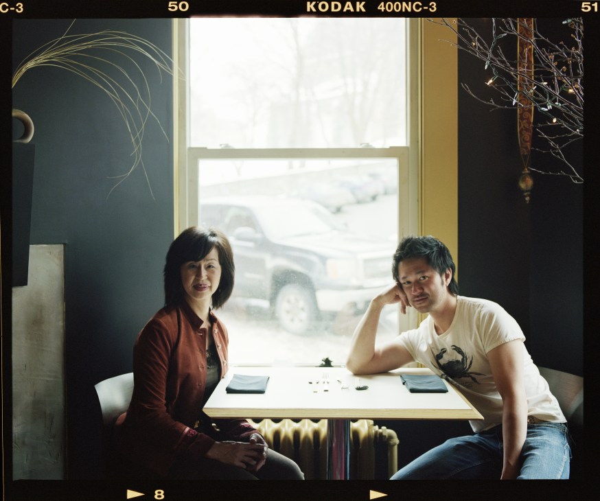 Chef Tak Ishiwata and his mother Fumiko Ishiwata sit at a white table in their restaurant Basho, with snowy landscape visible through the window behind them.