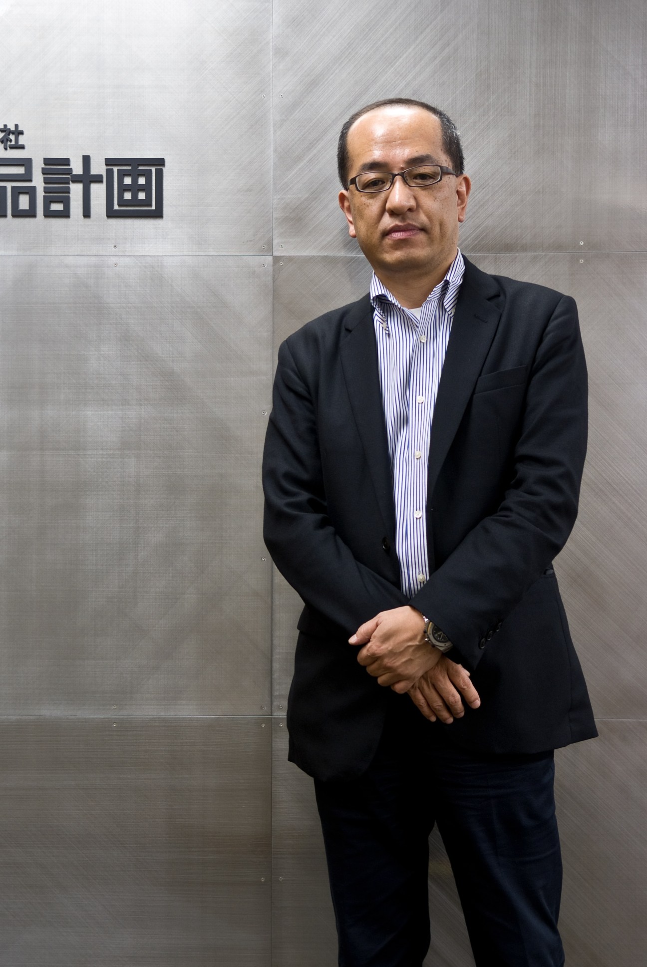 Takashi Yajima, general manager of Muji's Planning and Design Office, stands in business attire against a textured wall.