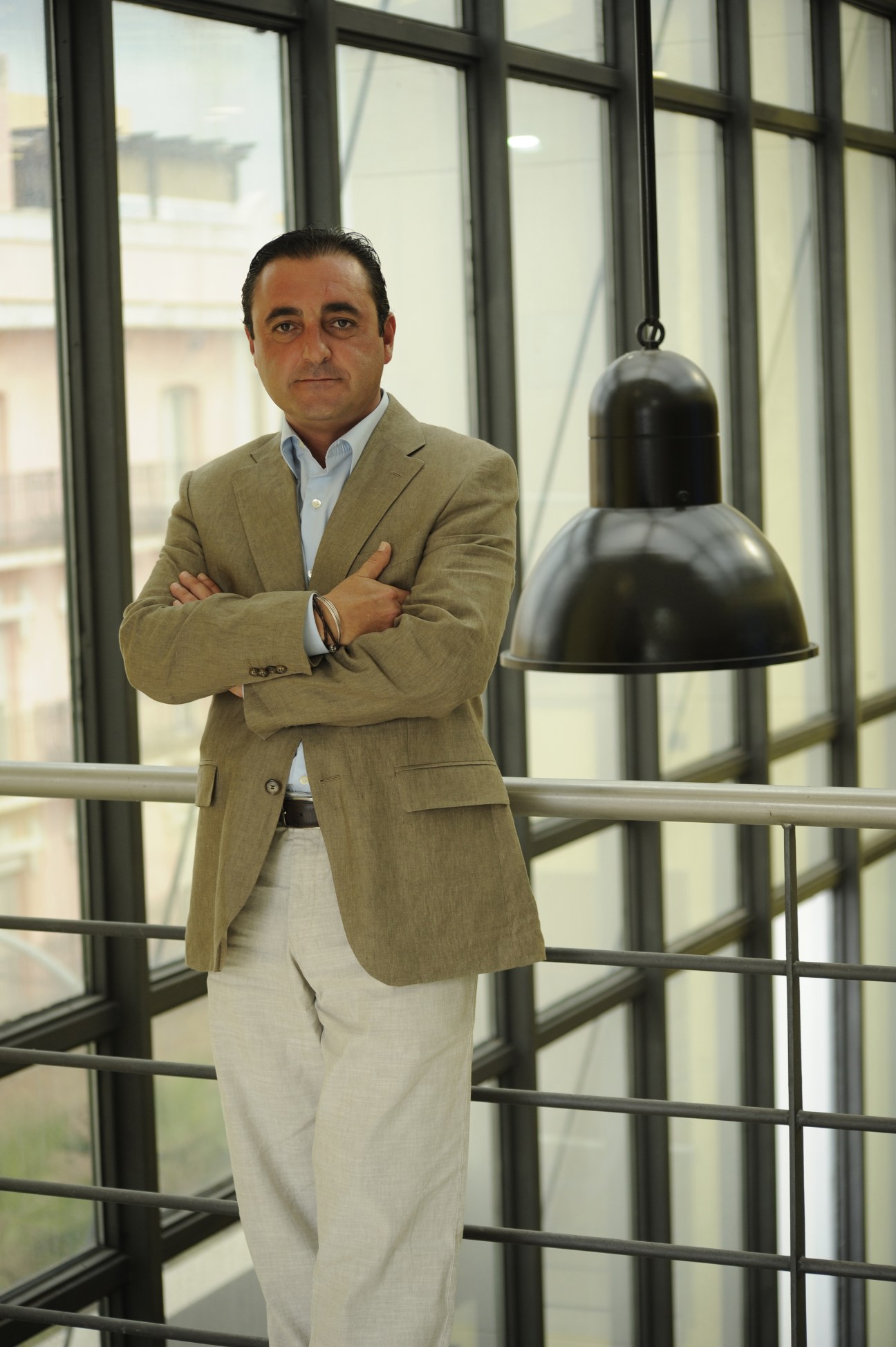 Man in beige blazer and white trousers stands with arms crossed by large windows in modern interior.