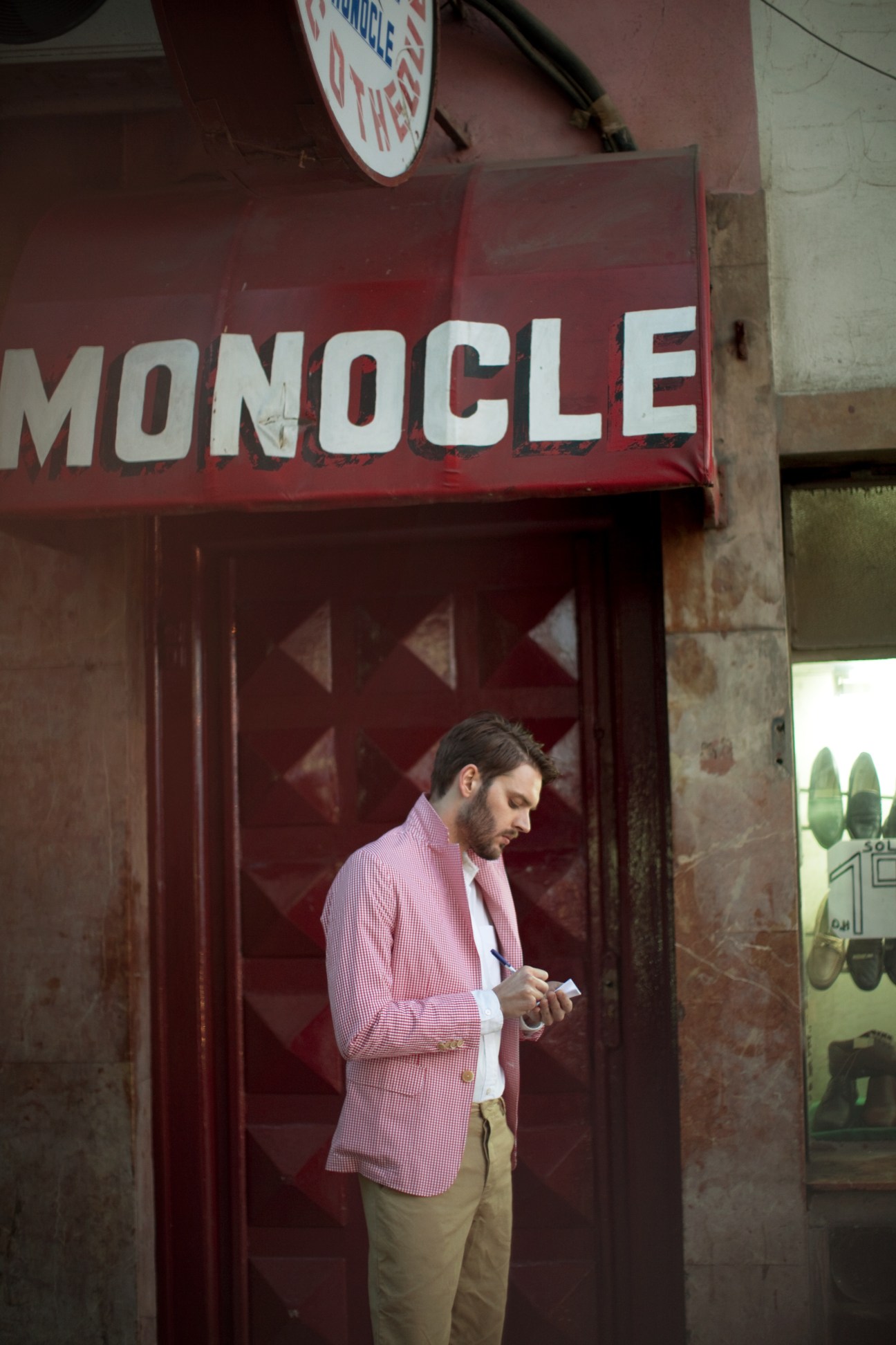 Man in pink jacket and light trousers standing in front of a red Monocle storefront door in Tangier.