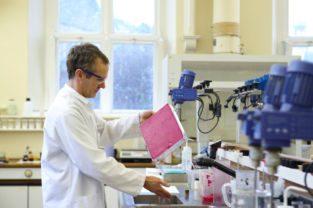 Technician in white lab coat holding pink paper sample in James Cropper laboratory with blue equipment.