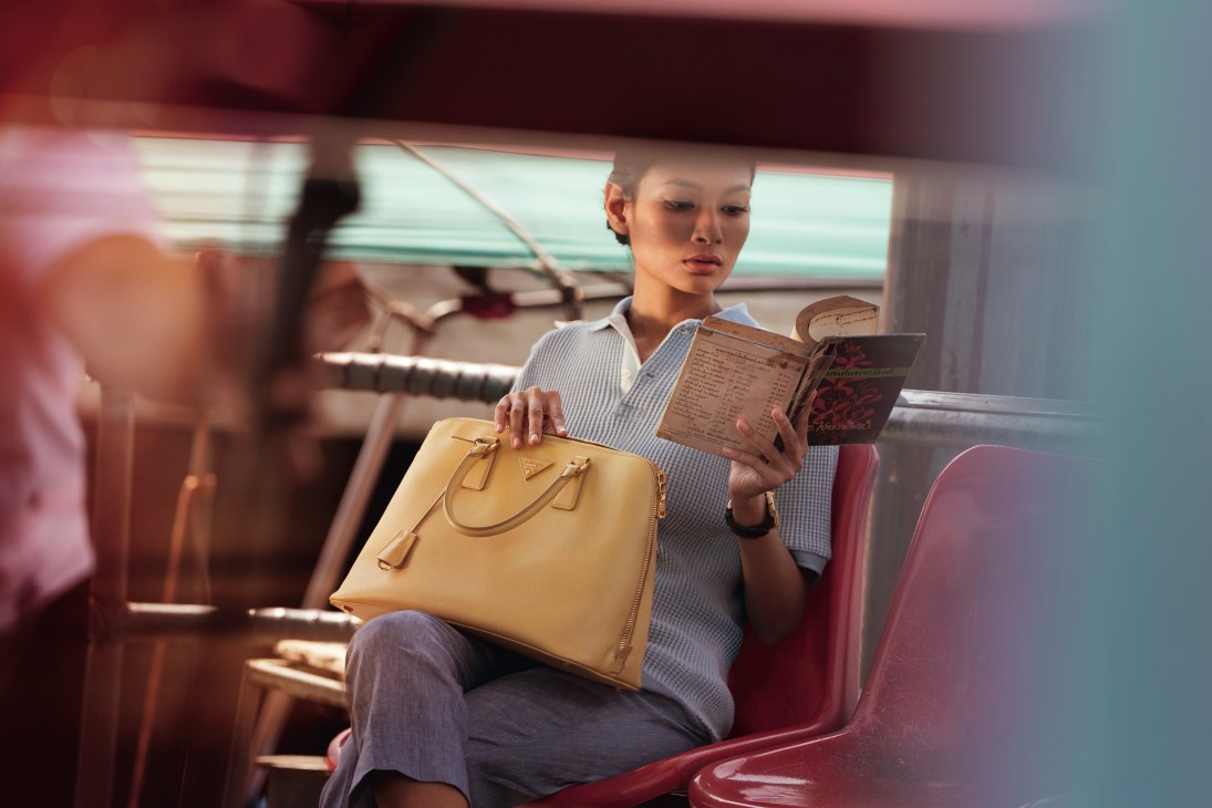 Woman in light blue shirt holding a yellow Prada bag and menu on a red seat in Bangkok.